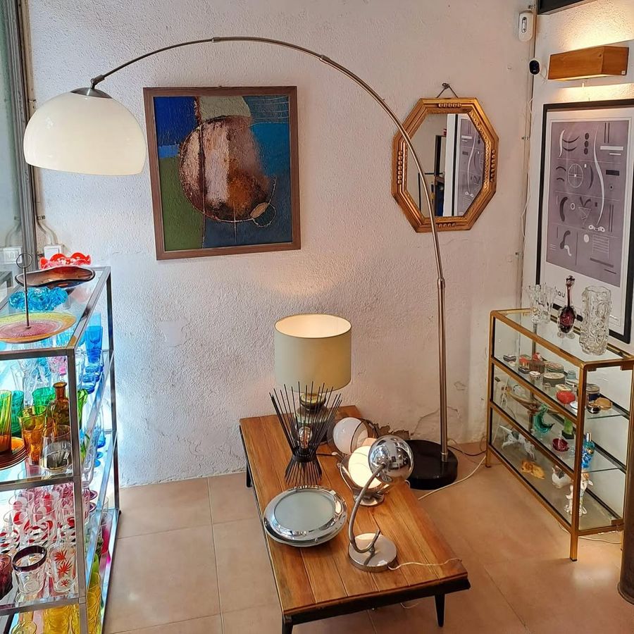 Interior de la botiga: un llum d’arc, una tauleta baixa de fusta amb llums de sobretaula, quadres i vitrines amb cristalleria de colors
